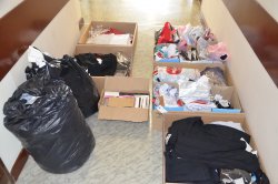 alt="Na korytarzu komendy Policji w Turku ustawione pięć kartonów z odzieżą, karton perfum oraz dwa czarnymi worki z ubraniami. Mienie zabezpieczone przez policjantów na targowisku."