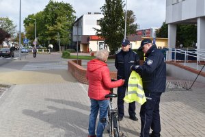 alt=&quot;Policjanci w trakcie przekazywania kamizelki odblaskowej rowerzystce przed budynkiem KPP w Turku.&quot;