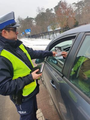 alt=&quot; Policjant w trakcie kontroli drogowej przekazuje ulotkę kontrolowanej osobie w samochodzie.&quot;