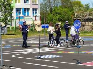 alt=&quot; Na zdjęciu policjant uniesioną dłonią, dwójka dzieci na rowerach w trakcie jazdy na torze.&quot;