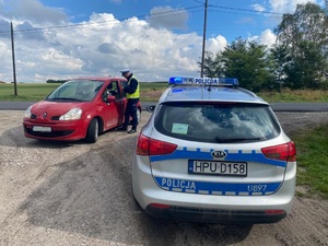 alt=&quot;Policjant podczas kontroli samochodu marki Renault Clio koloru czerwonego. W pierwszym planie po prawej stronie radiowóz policyjny Kia&quot;.
